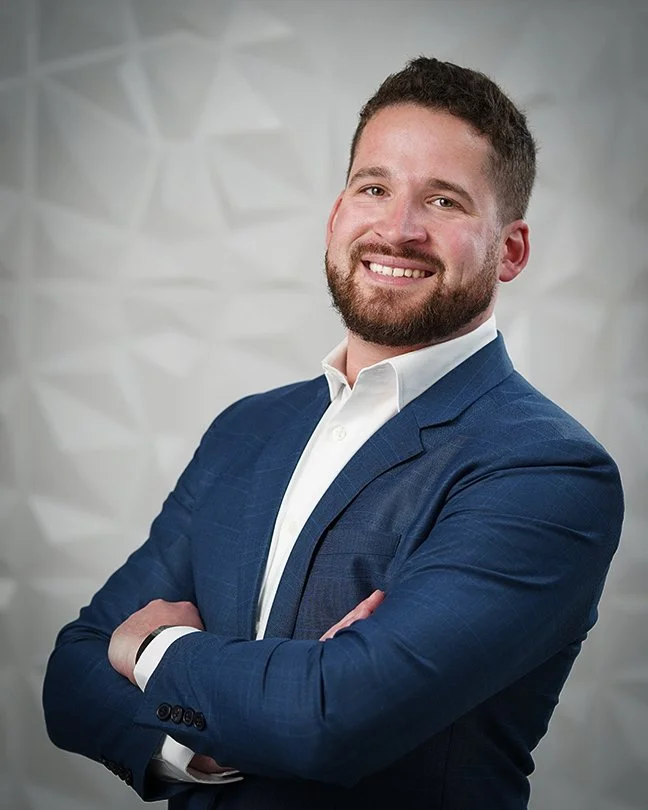 Portrait of a smiling man with a beard in a blue suit and white shirt, crossing his arms, standing in front of a textured light gray wall.