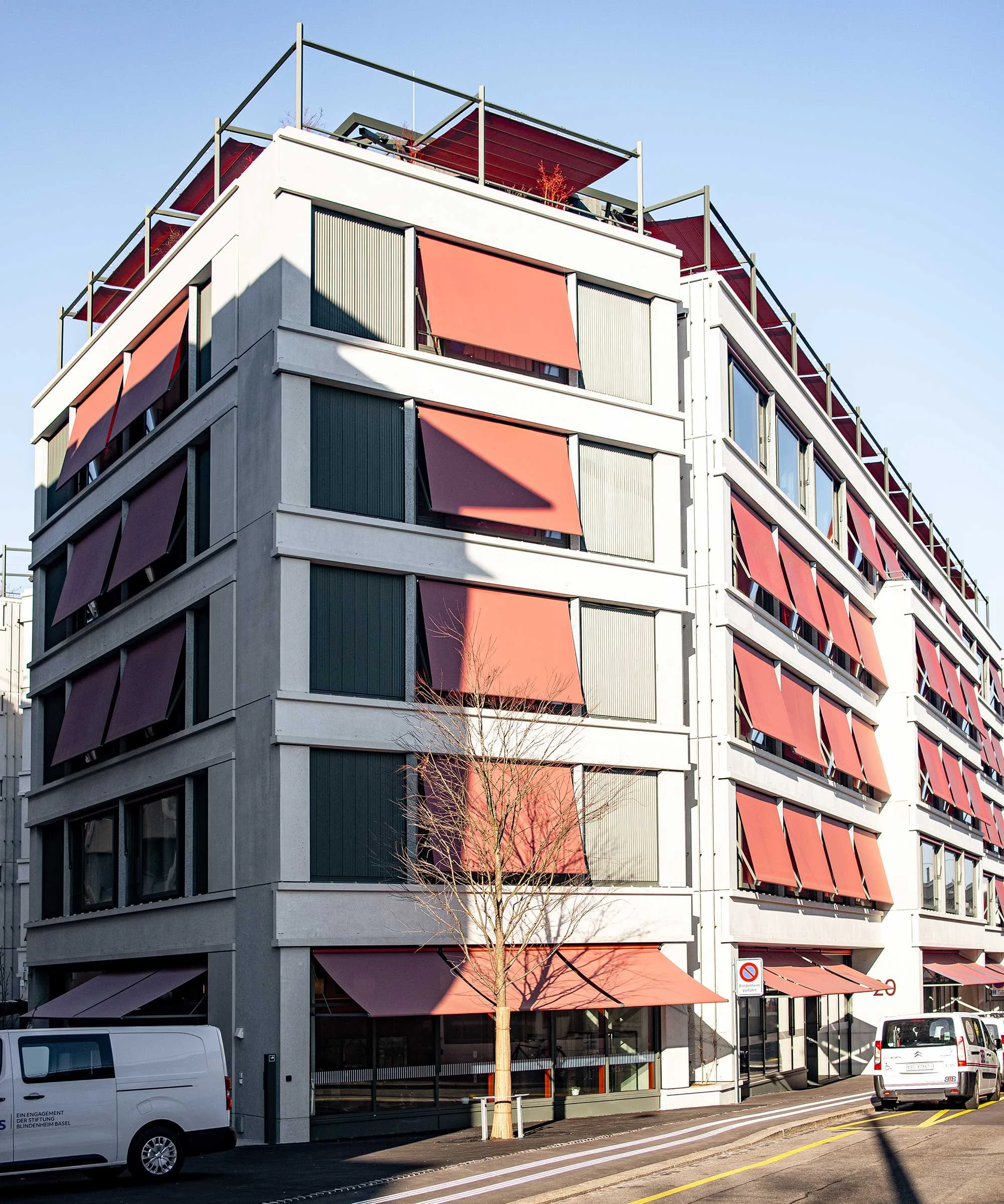 Modernes mehrstöckiges Wohngebäude mit Balkonen und begrüntem Dach, Straßenszene mit Fußgängern, Bäumen und Autos, abends.