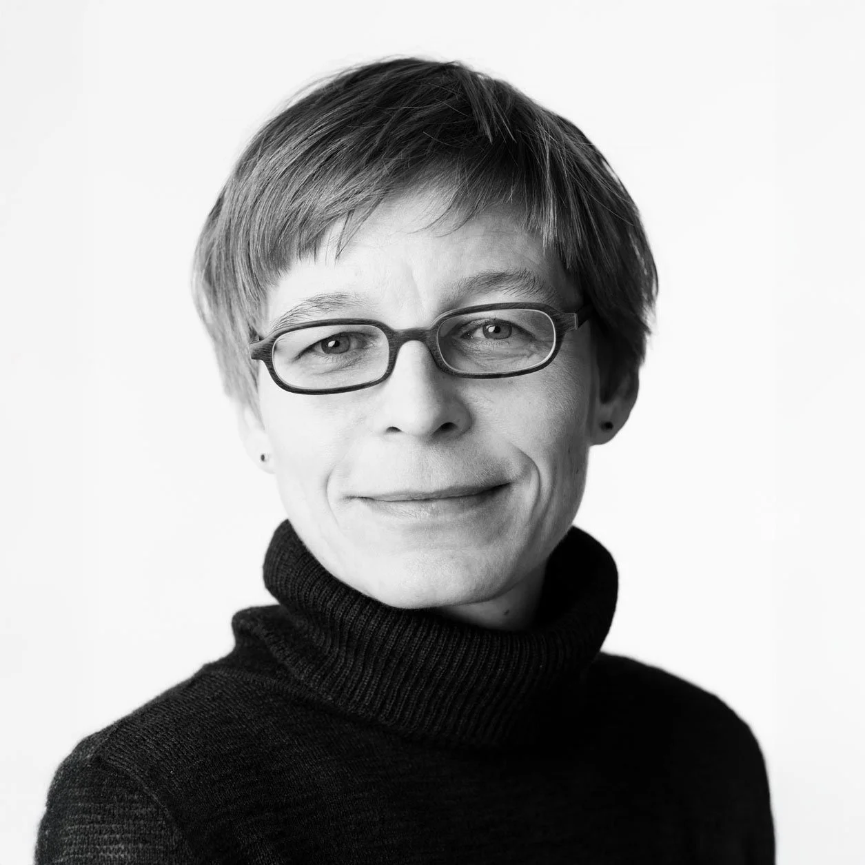 Schwarz-weiße Porträtaufnahme einer Frau mit kurzen Haaren, Brille und dunklem Rollkragenpullover, lächelnd