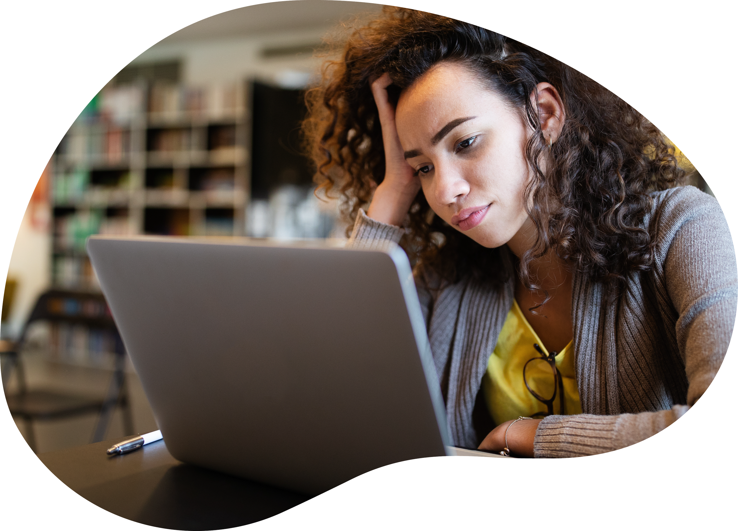 A woman with curly hair looking at a laptop with a worried or stressed expression, resting her head on her hand, in a library or bookstore.