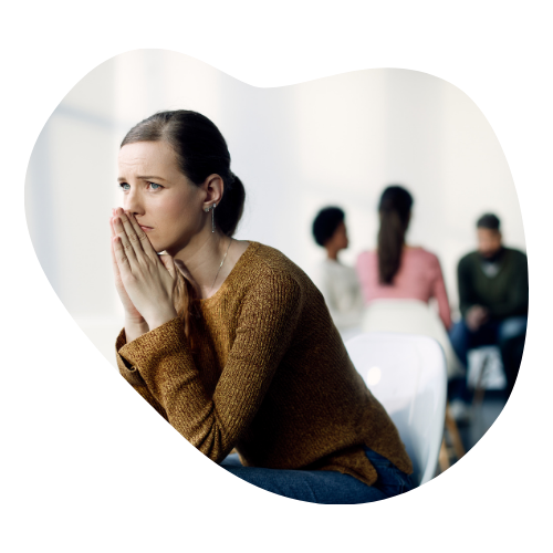 A woman sits apart from a group, hands clasped at her mouth with a worried expression, representing feeling like an outsider with ADHD.