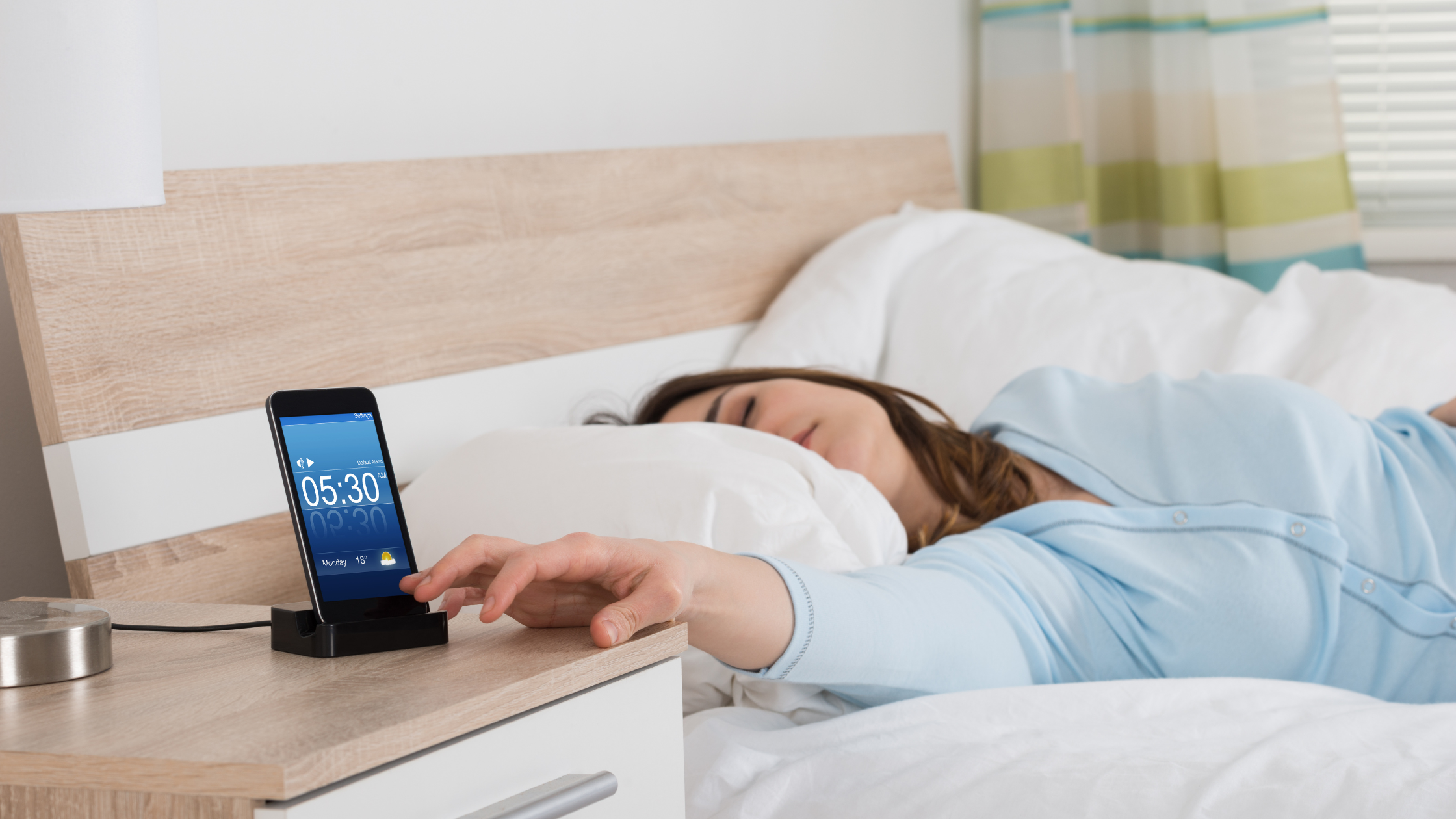 Woman sleeping in bed, reaching to silence a 5:30 AM alarm on her nightstand.