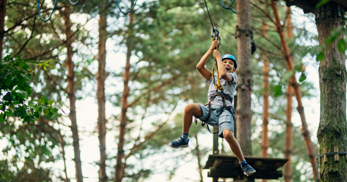 A smiling boy in a blue helmet rides a zipline through tall trees, conveying the thrill of taking a leap of faith.