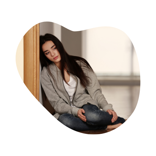 A young woman sits alone on the floor leaning against a wall, looking sad and withdrawn.