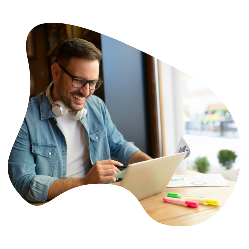 A man wearing glasses sits smiling working on a laptop. He has headphones around his neck and he is sitting in a coffee shop.