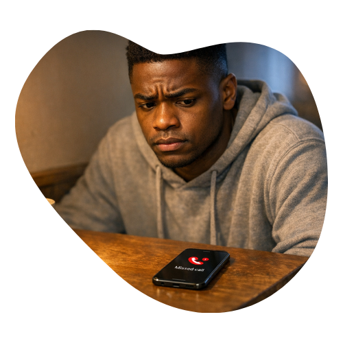 A man in a gray hoodie sits at a table looking distressed at a phone displaying a missed call notification — representing the social withdrawal and avoidance that can accompany ADHD masking.