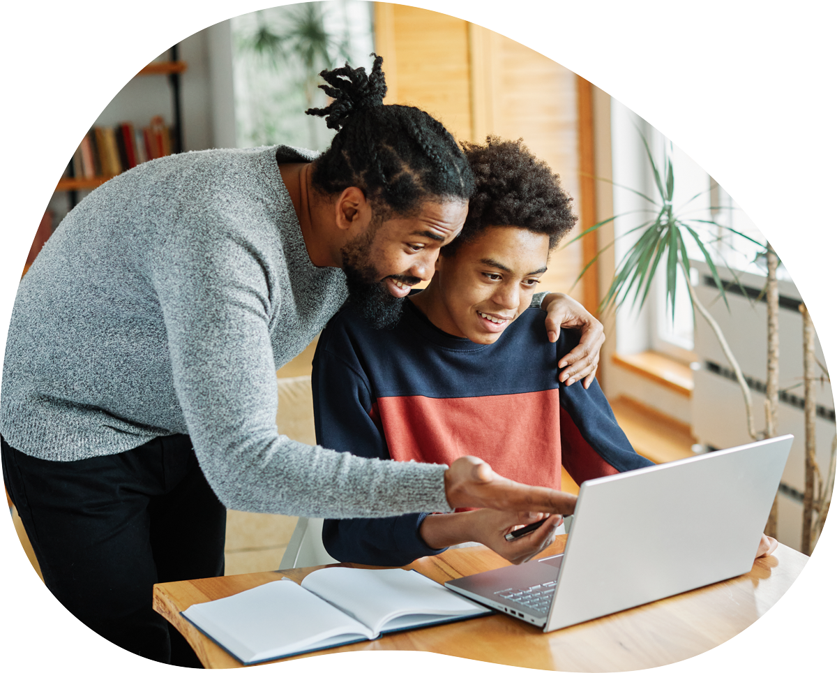 A man and boy look at a laptop screen together in a bright, cozy room with a bookshelf and potted plant.