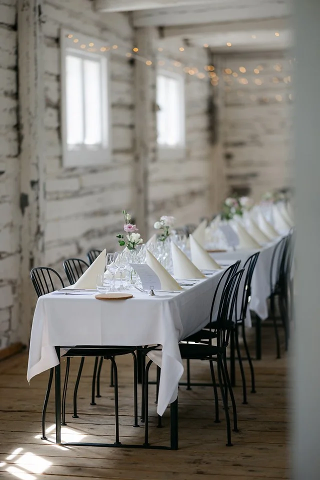 Elegant dining table set with white tablecloth, folded napkins, wine glasses, floral centerpieces, and black chairs in a rustic venue with wooden walls and string lights