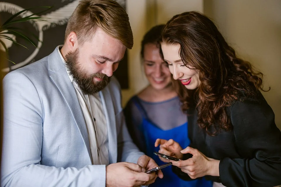 Three people, two women and a man, are smiling and looking at their smartphones together in an indoor setting.