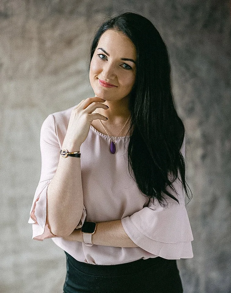 A woman with long black hair smiling, wearing a light pink blouse, a purple pendant necklace, a smartwatch, and a bracelet, standing against a neutral background.