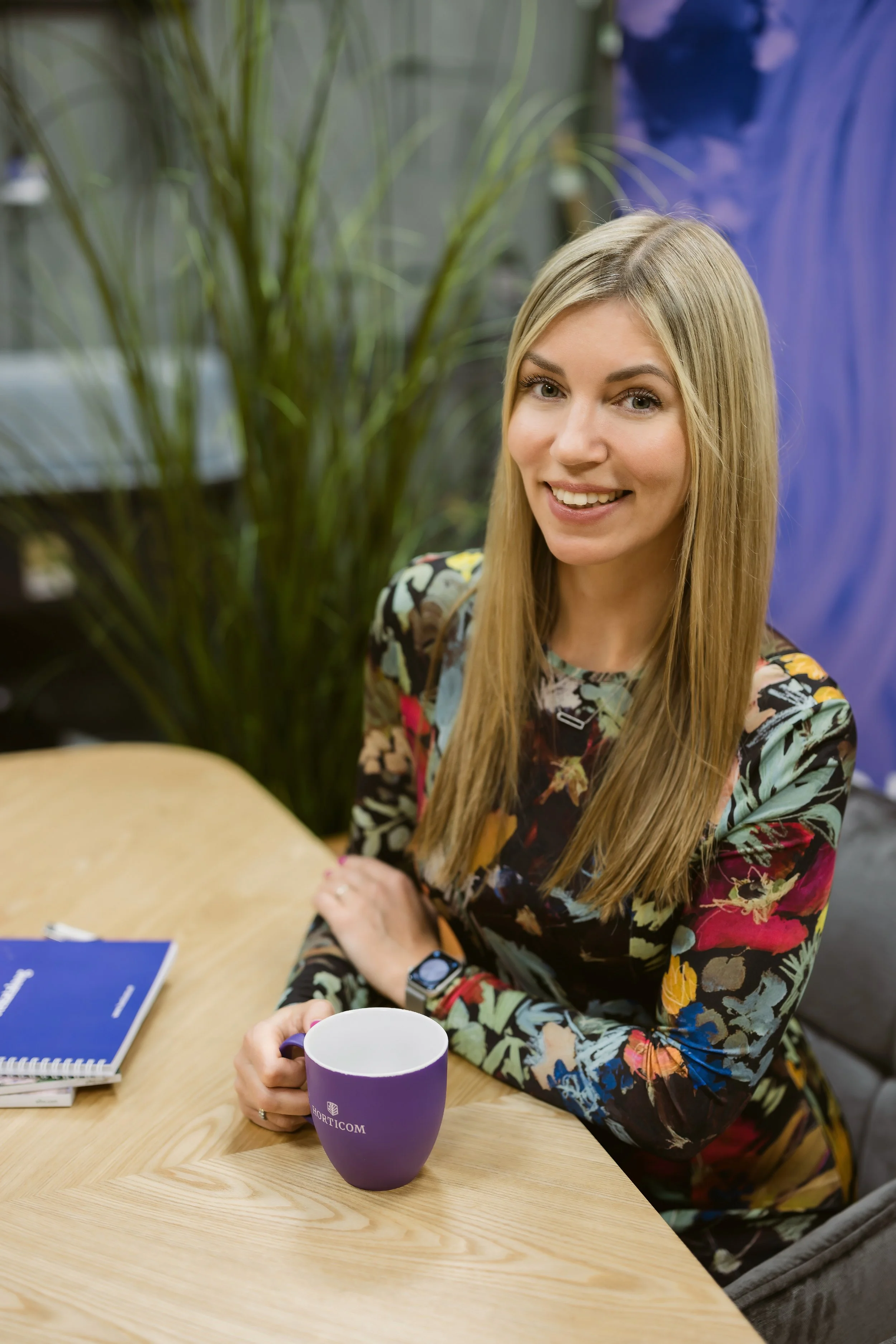 A woman with long blonde hair sitting at a wooden table, holding a purple mug, smiling at the camera. She is wearing a colorful floral patterned top, with a book or notebook and a smartwatch on her wrist. There is a plant and some colorful fabric or 