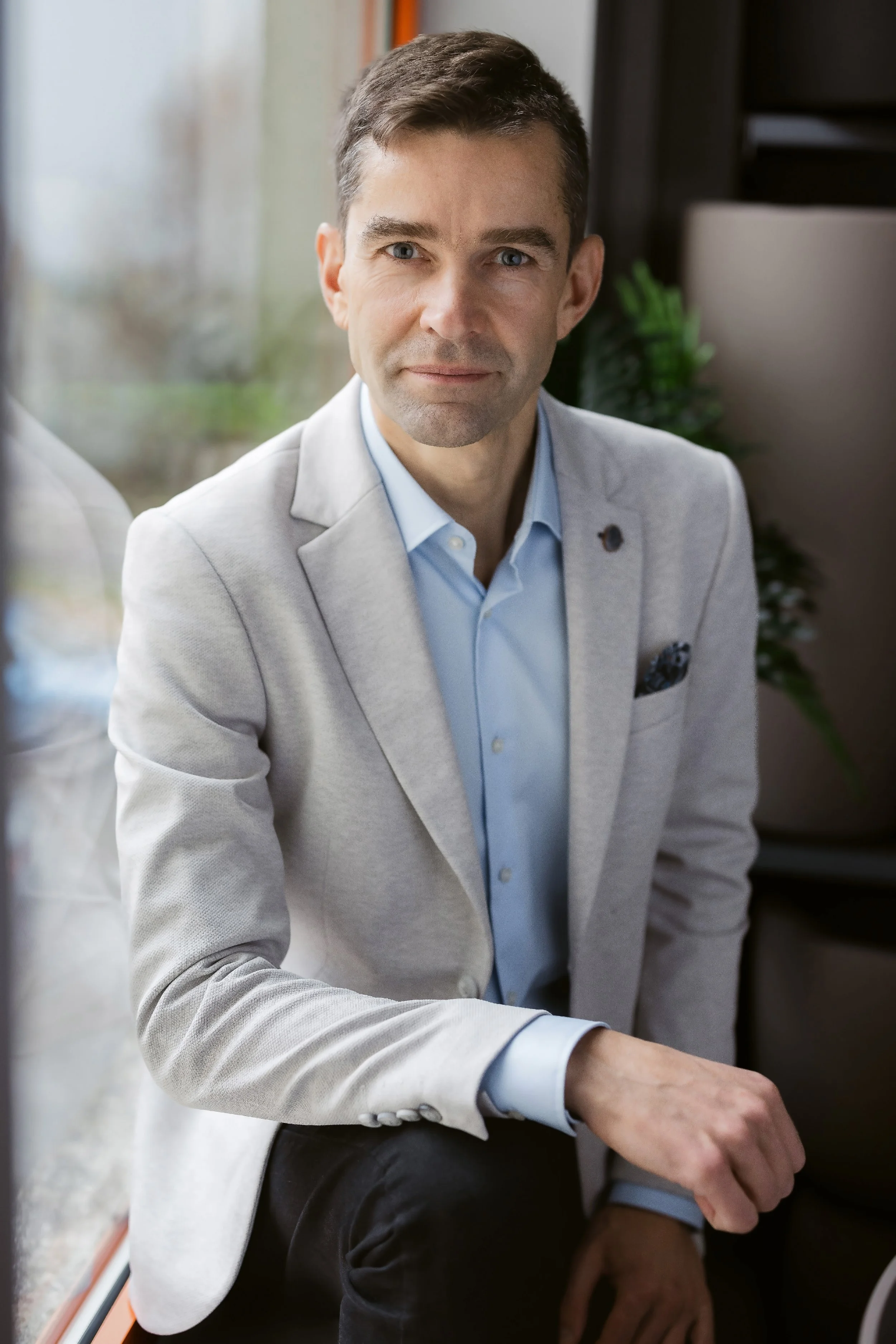 A man in a light gray suit and blue shirt sitting indoors near a window.