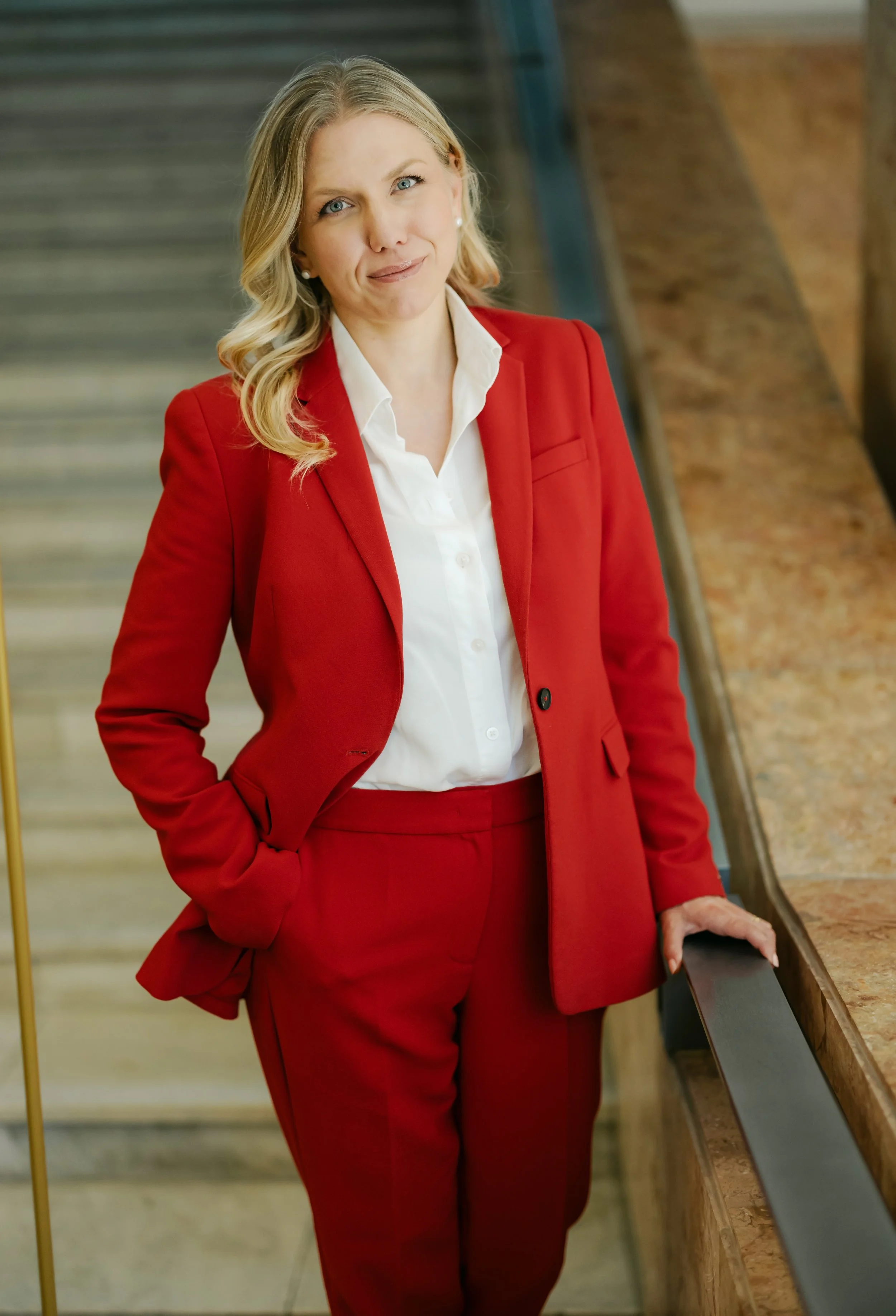 A woman with blonde hair, wearing a red suit and white shirt, standing on a staircase with her left hand resting on a marble railing.