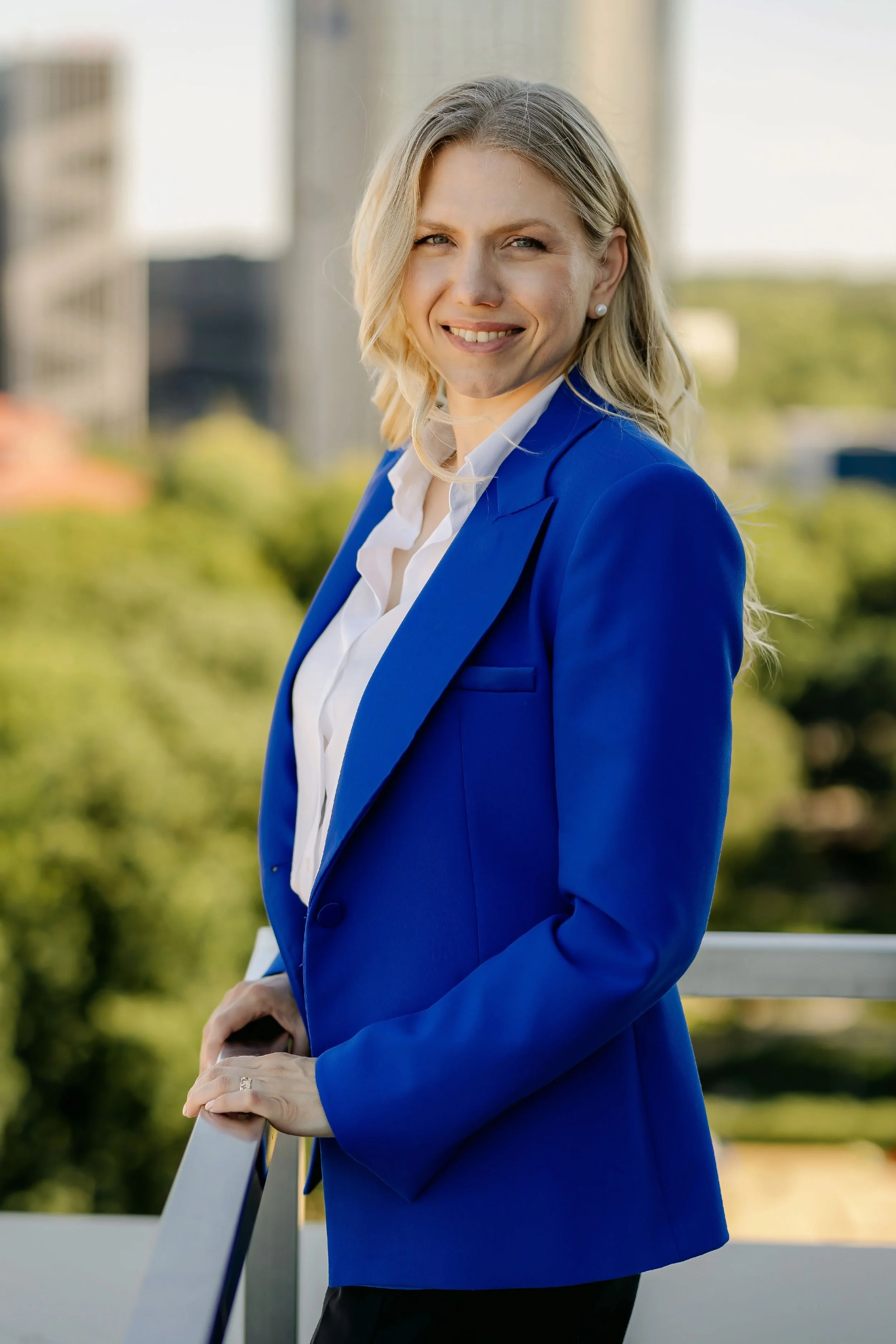 A woman in a blue blazer and white shirt standing outdoors on a sunny day, leaning on a railing with a cityscape in the background.