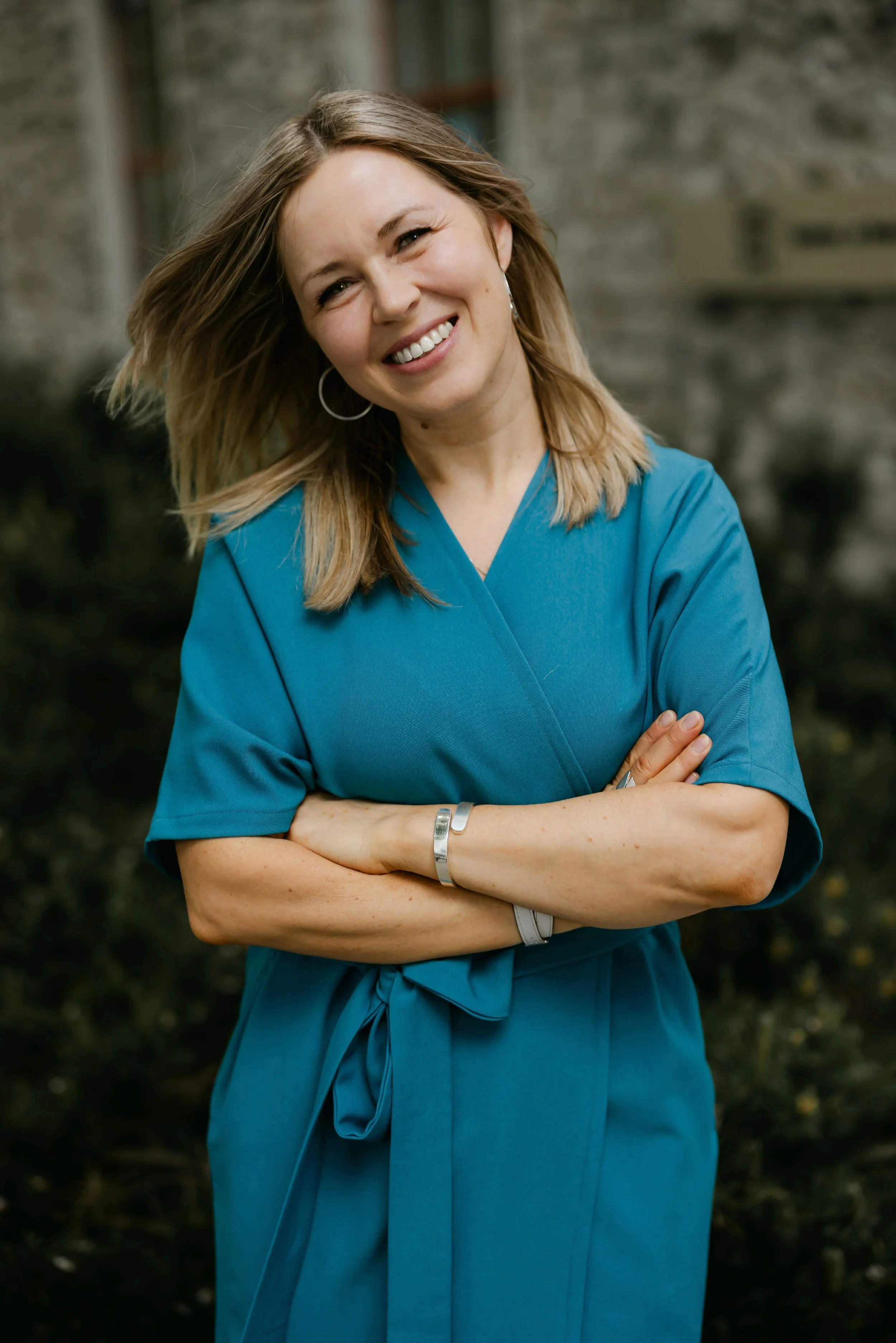 Smiling woman with blonde hair wearing teal scrubs, standing outdoors with crossed arms.