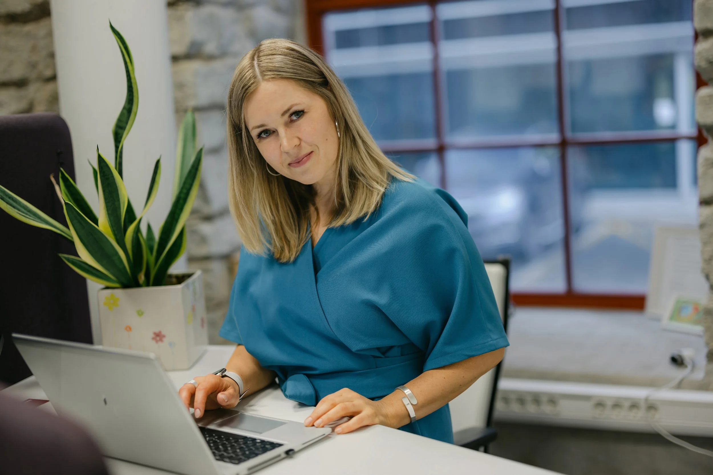 A woman in a blue uniform working on a laptop in an office with a potted plant and a brick wall.