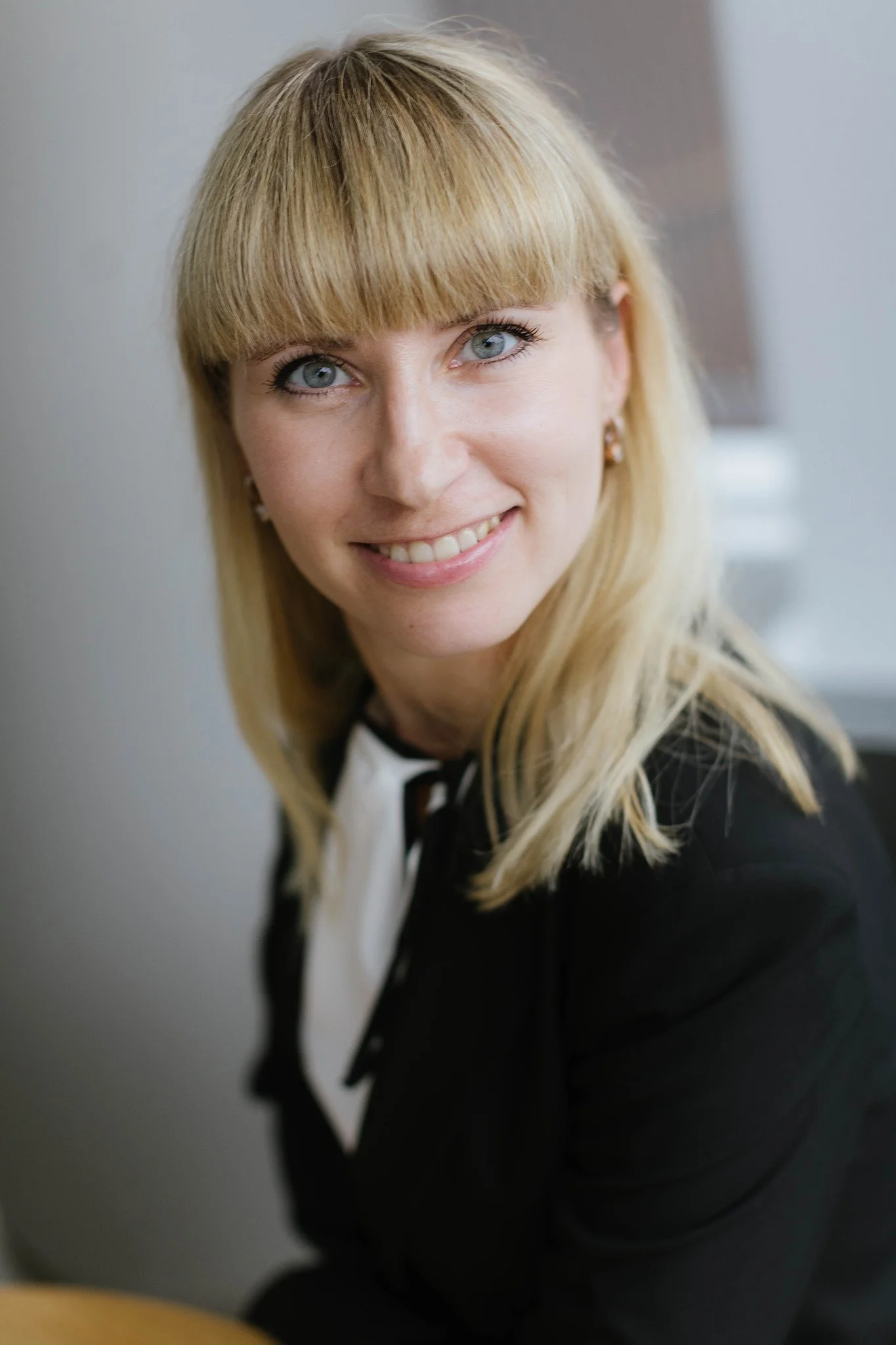 Close-up of a smiling woman with blonde hair and blue eyes, wearing a black blazer with a white blouse.