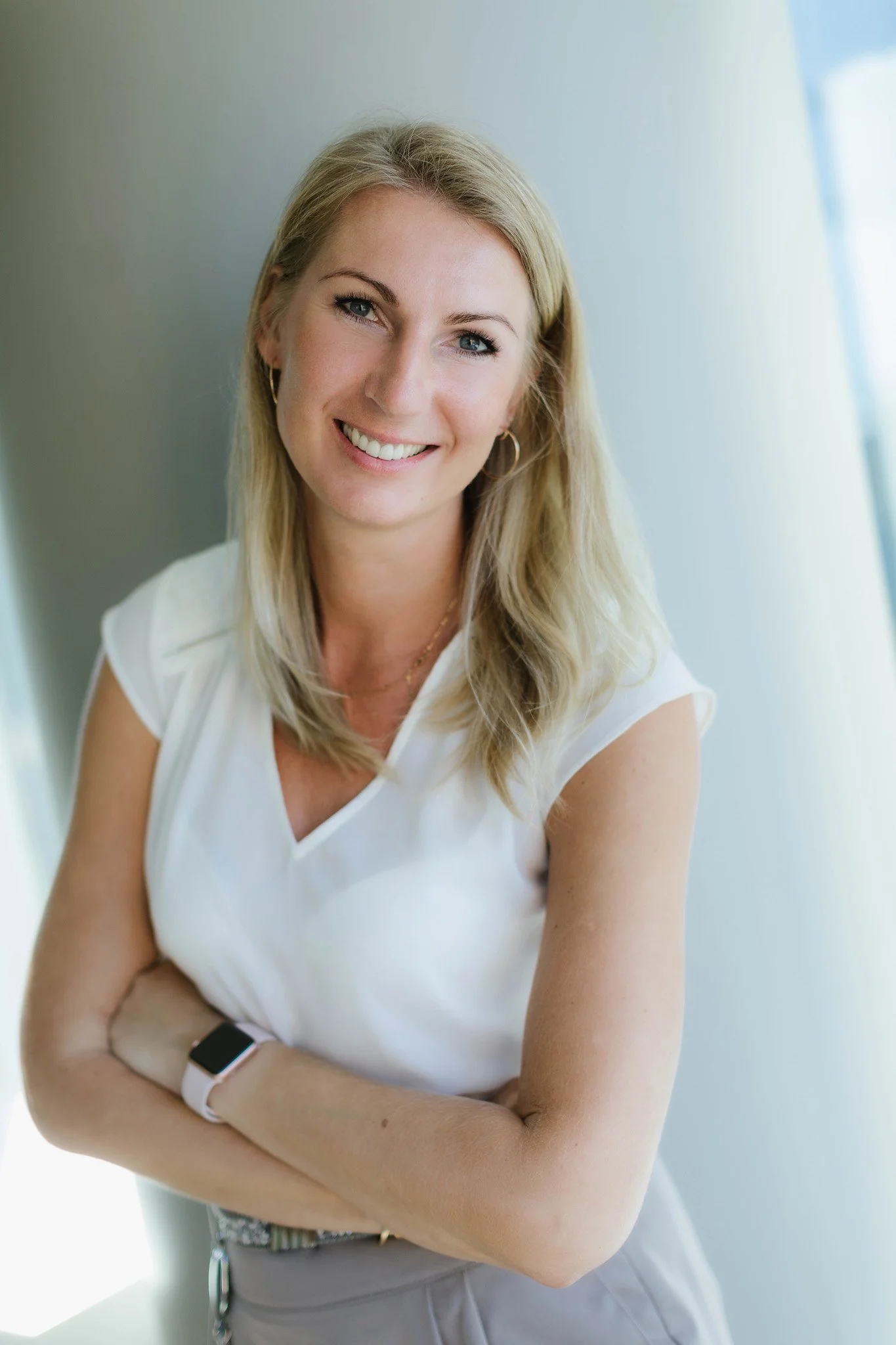 A smiling woman with blonde hair wearing a white top and a smartwatch, standing near a window with sunlight.
