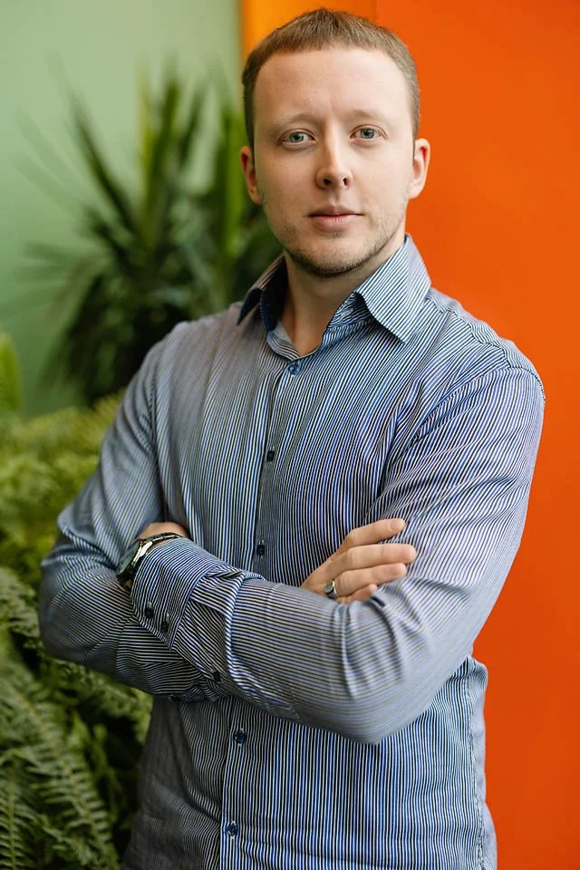A man with short hair in a blue striped shirt standing with crossed arms in front of a green plant and orange wall.