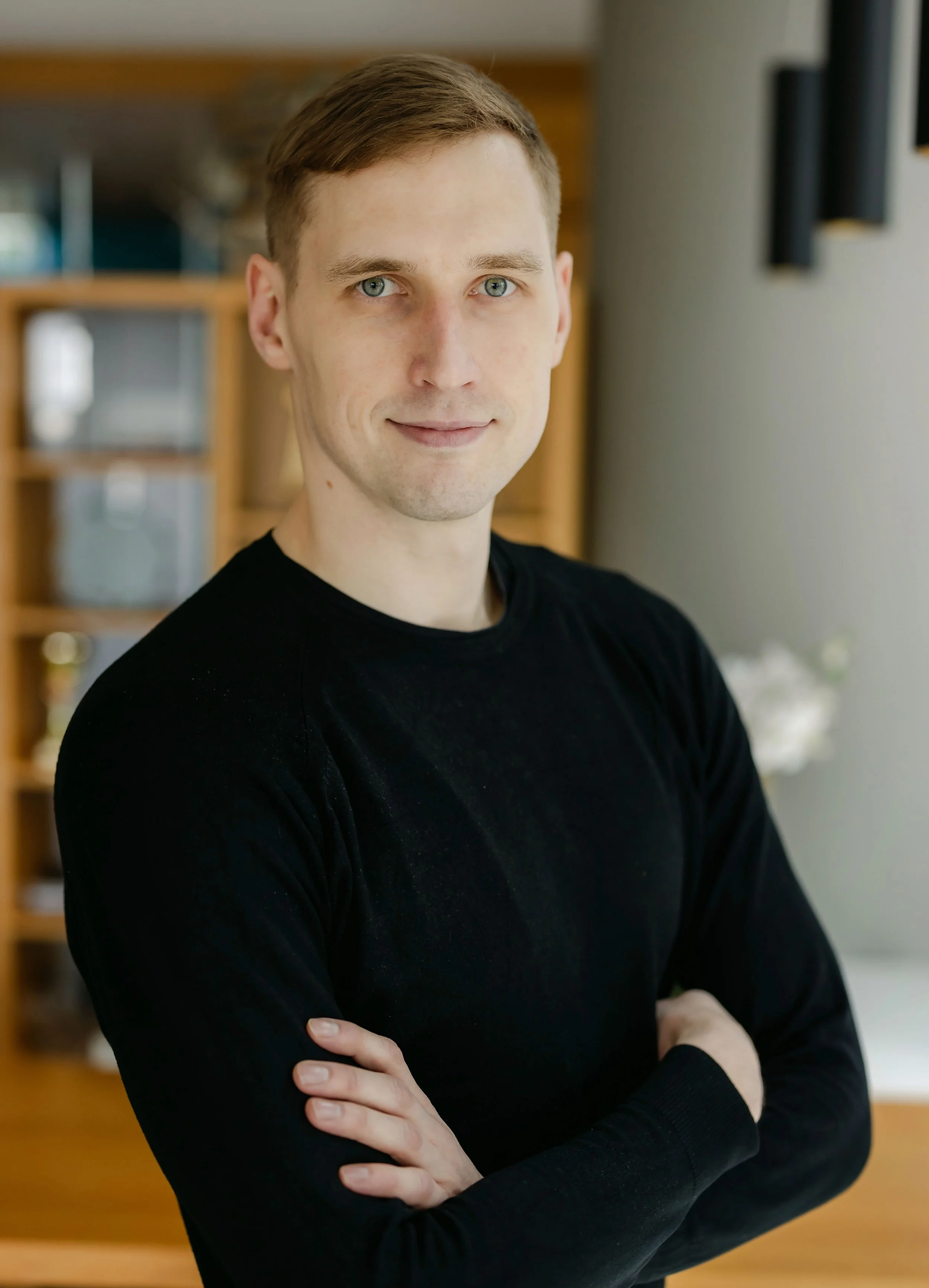 A young man with light skin, short light brown hair, and blue eyes, wearing a black long-sleeve shirt, standing with arms crossed in a well-lit room with wooden furniture and a flower arrangement in the background.