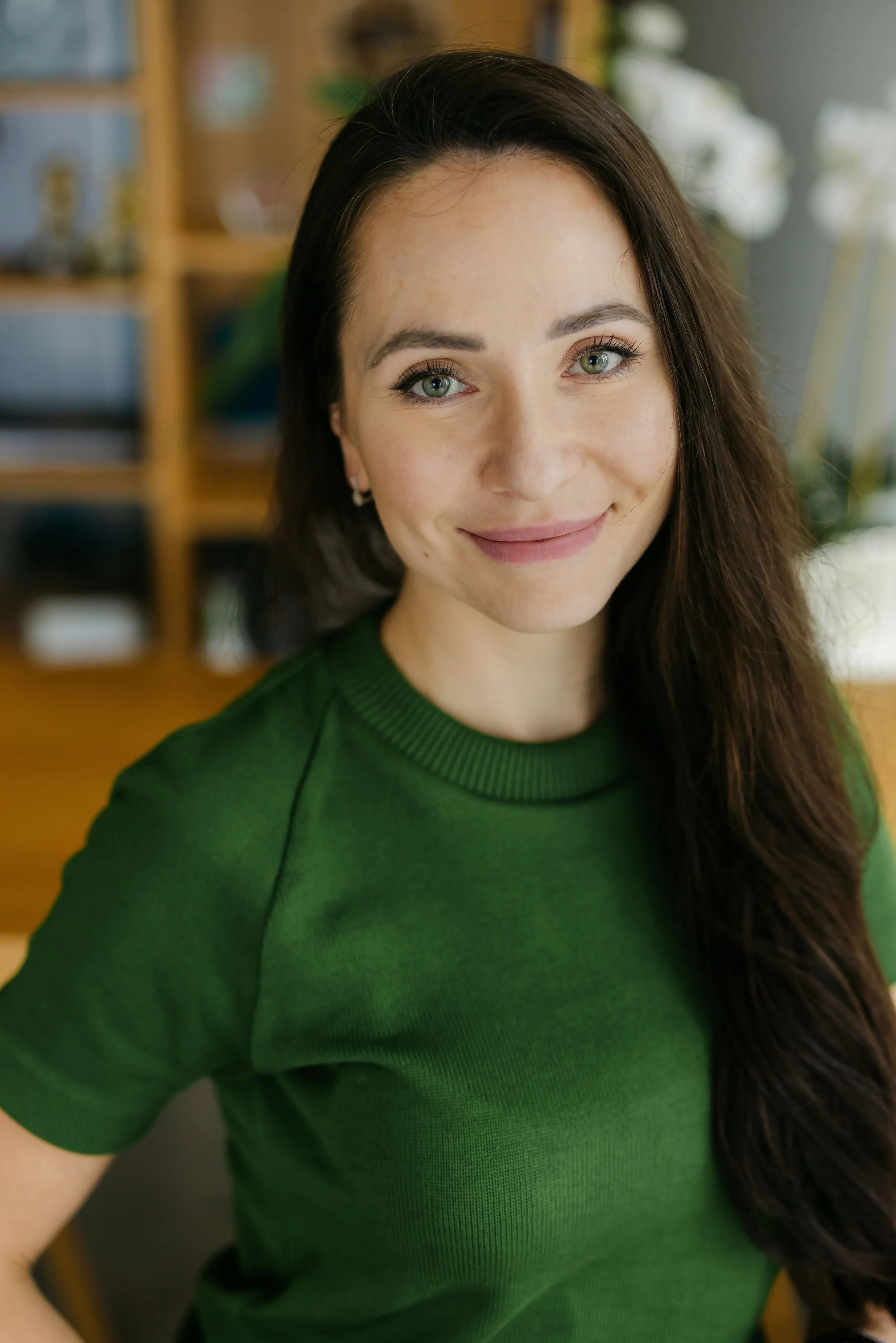 A woman with long dark hair in a green top smiling indoors.