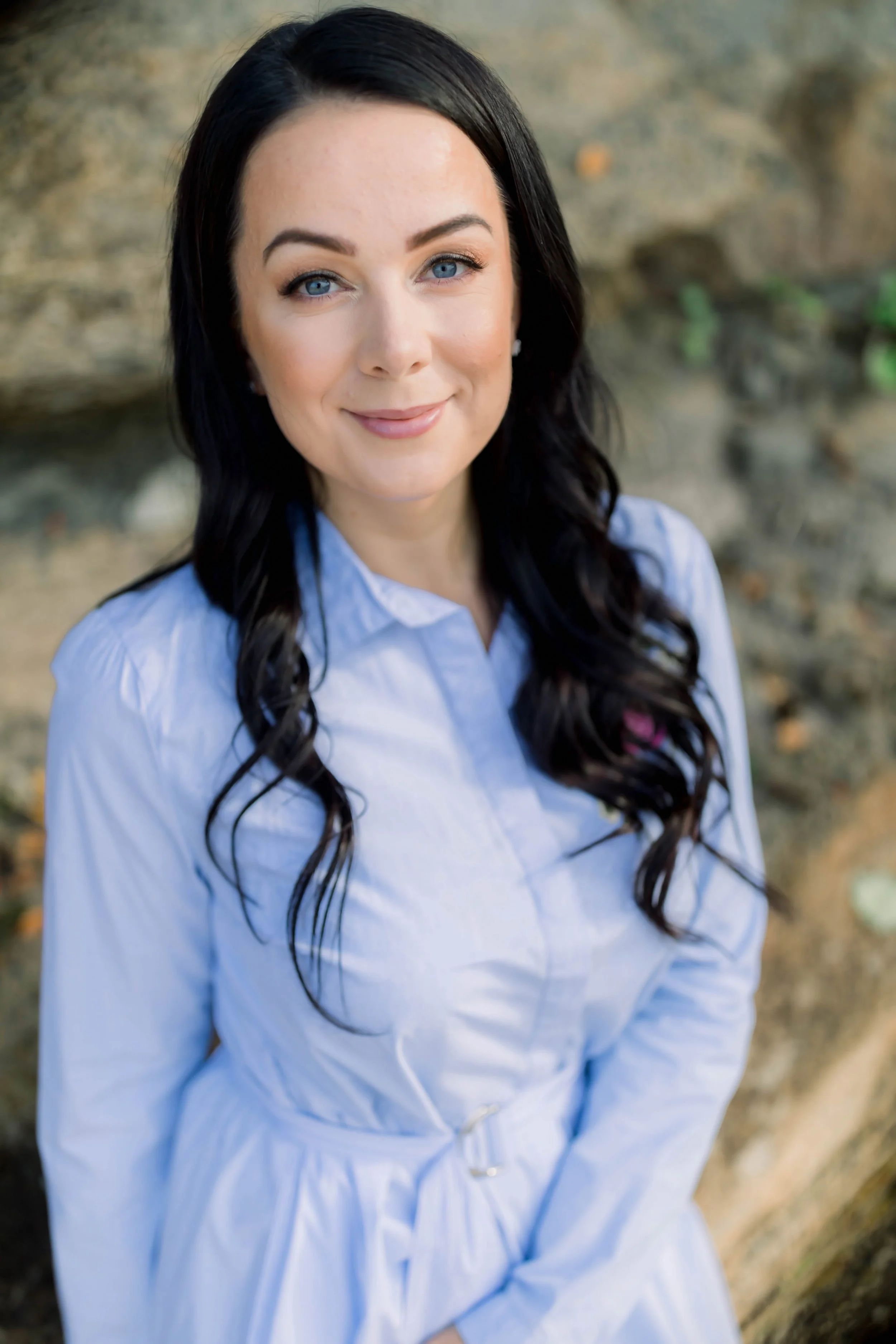 A woman with long dark wavy hair and blue eyes, wearing a light blue button-up shirt, standing outdoors near a tree trunk.
