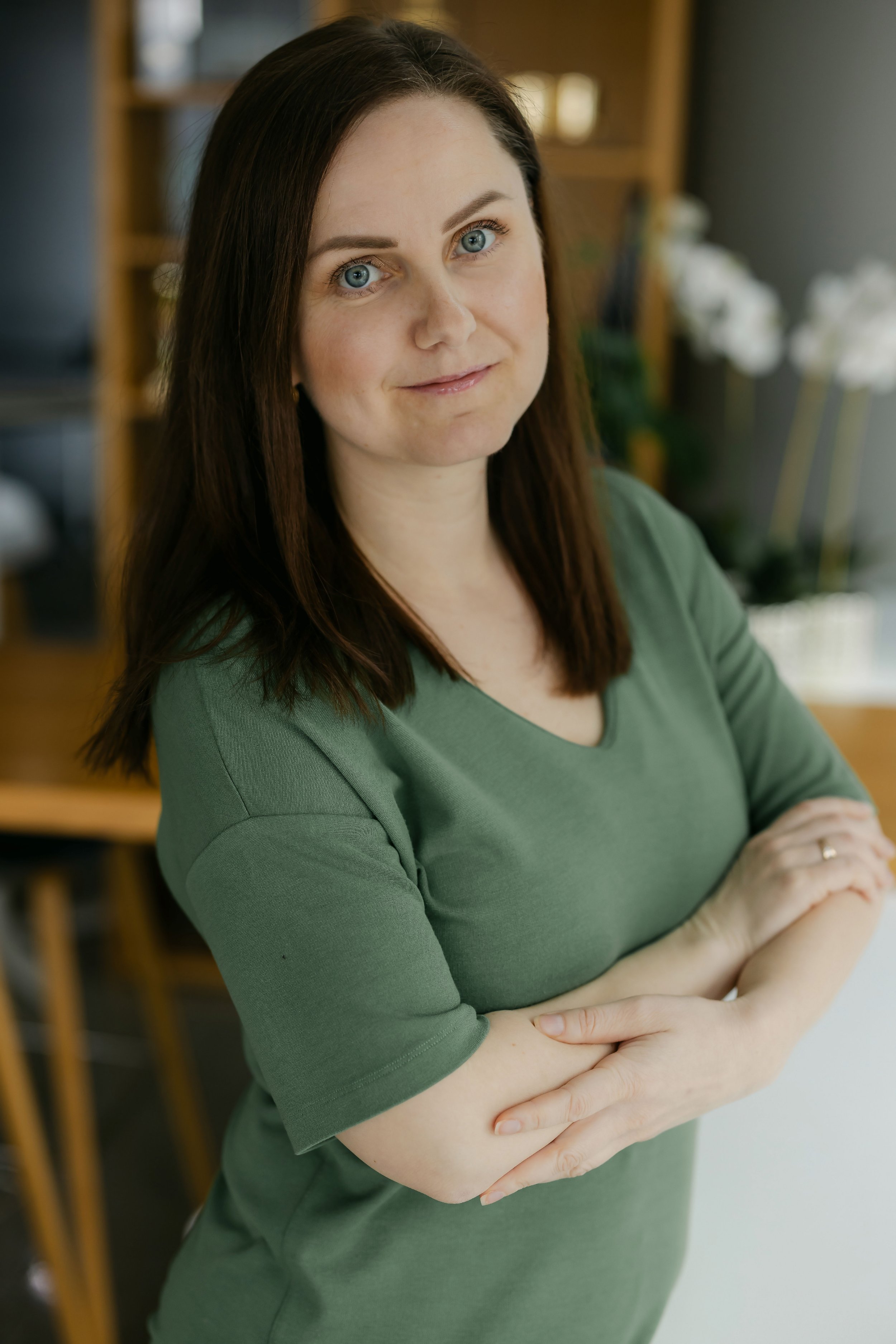 A woman with brown hair and blue eyes wearing a green top, standing indoors with arms crossed, in front of a background with blurred furniture and flowers.