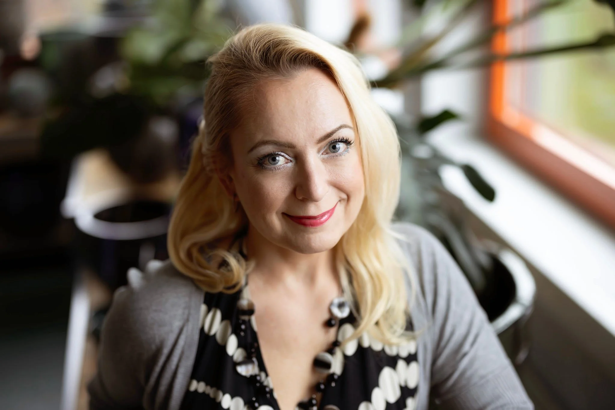A woman with blonde hair and blue eyes smiling and looking at the camera, in a brightly lit room with plants and a window.