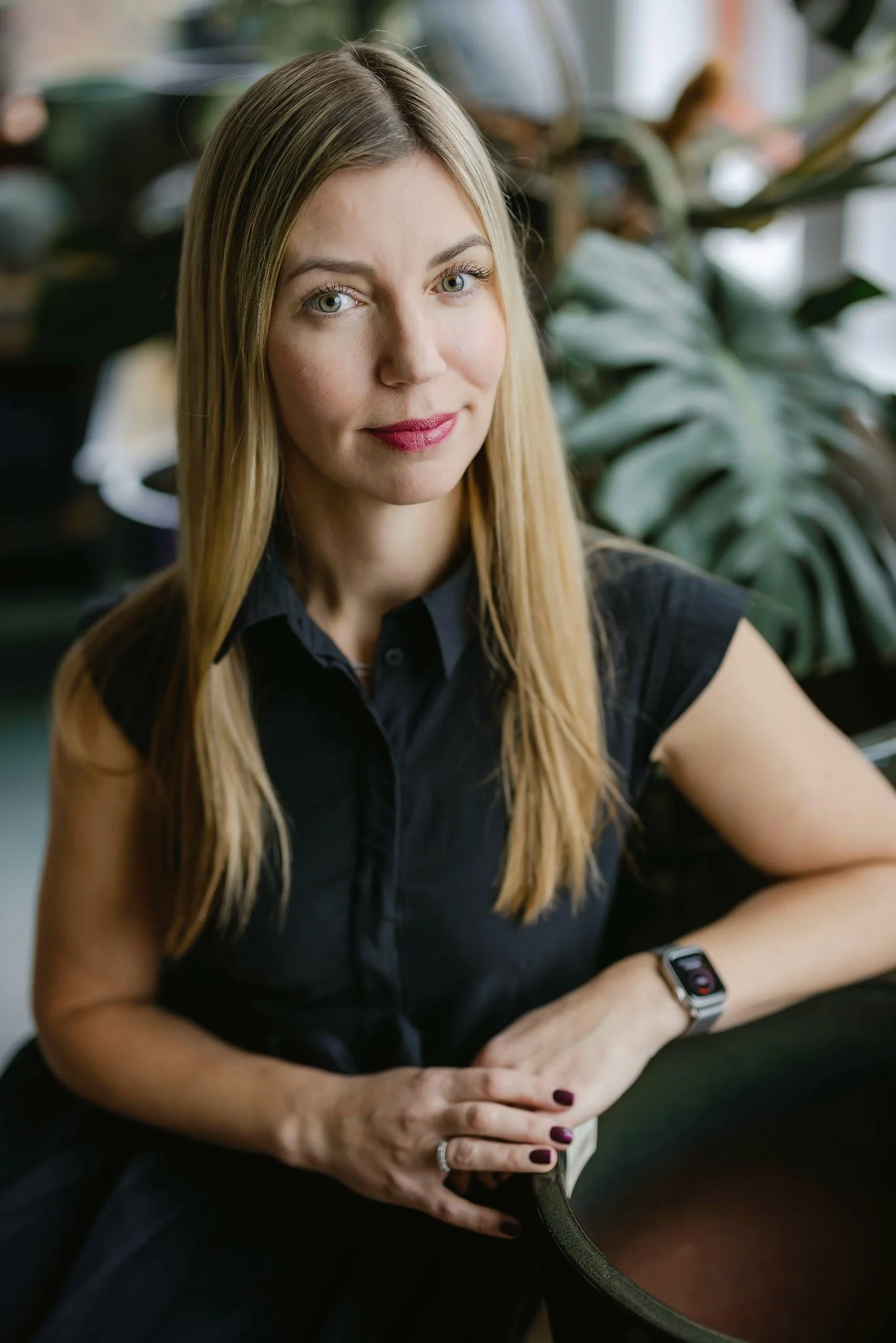 A woman with long blonde hair and blue eyes, wearing a black sleeveless shirt and a smartwatch, sitting at a table with green plants in the background.