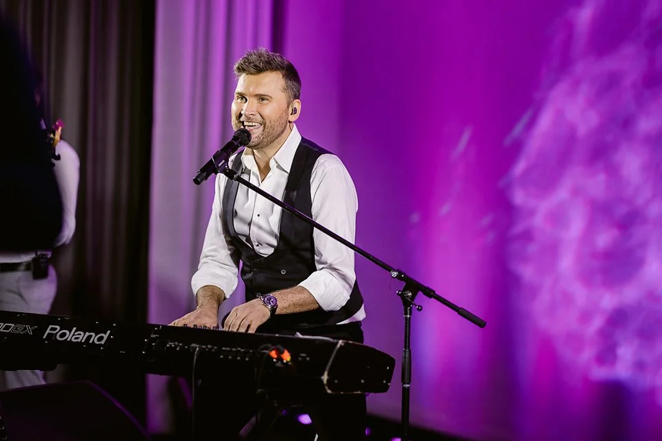 A man playing a keyboard and singing into a microphone during a live performance with a purple background.