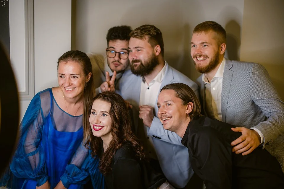 Group of six young adults taking a group photo indoors, smiling and making peace signs.