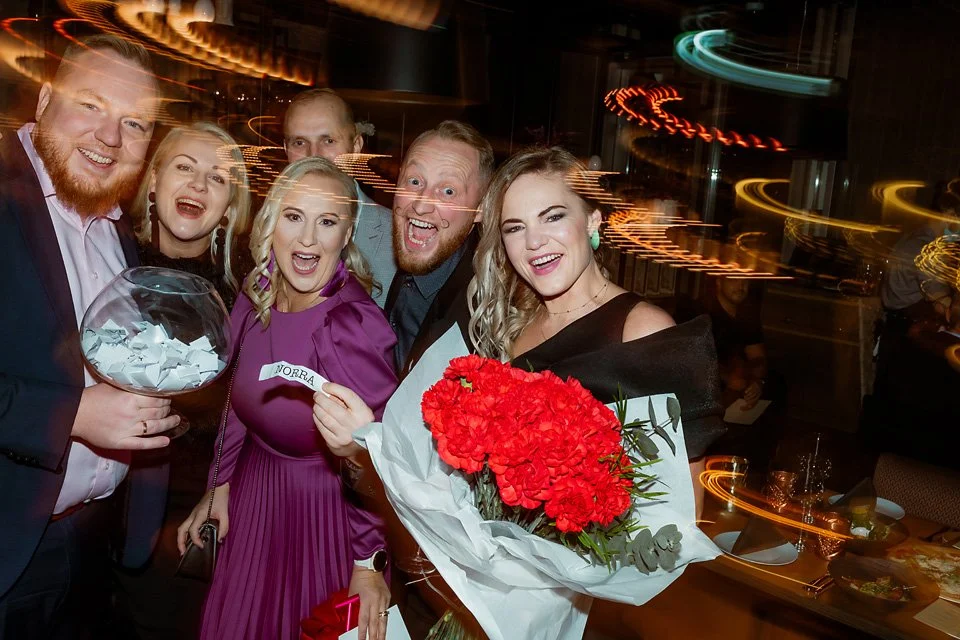 Group of six people celebrating at a party, smiling with one woman holding a large bouquet of red flowers and another holding a glass bowl with raffle tickets, in a dimly lit environment with light streaks in the background.
