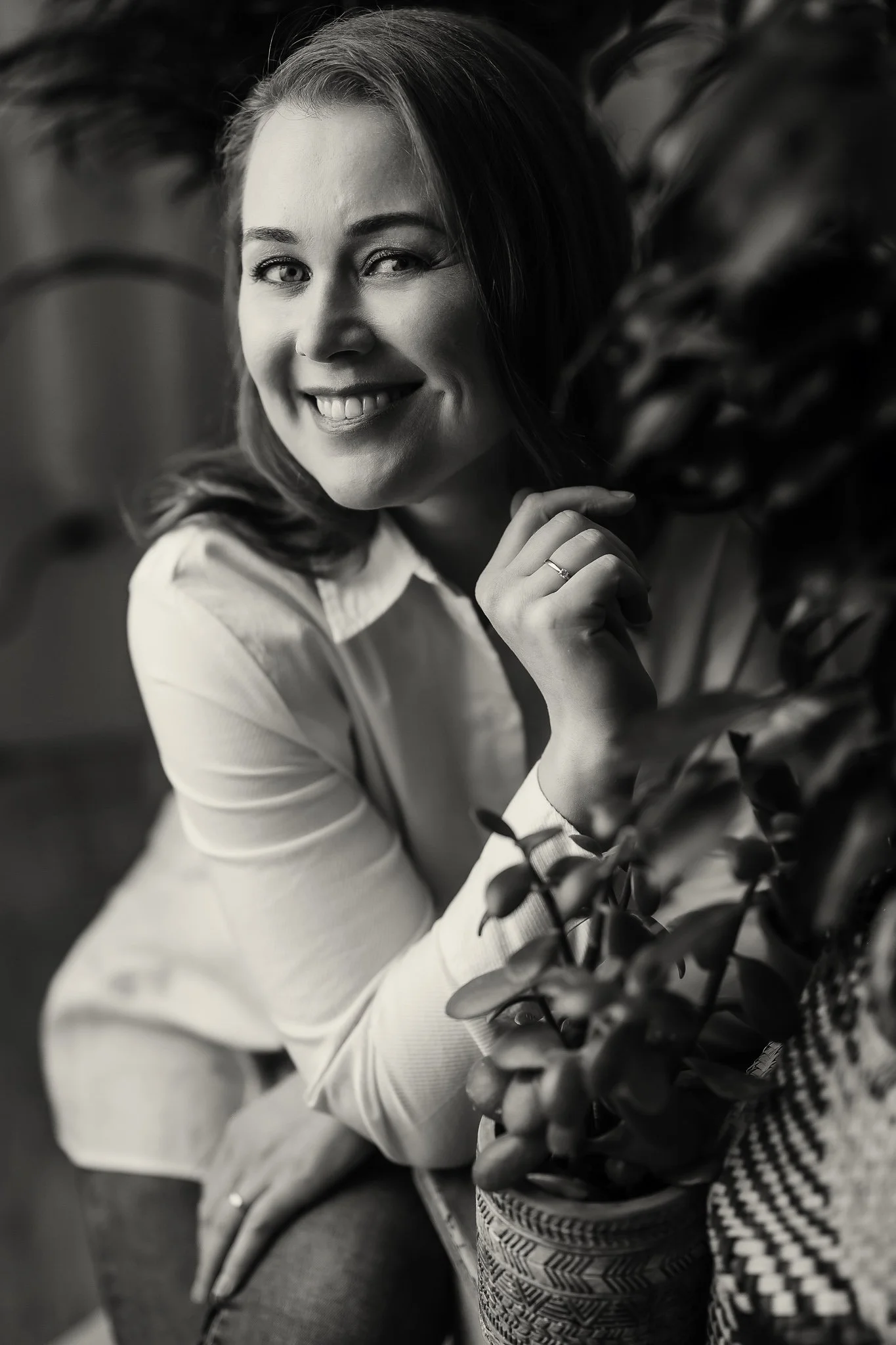 A woman with shoulder-length hair smiling and leaning on a potted plant.