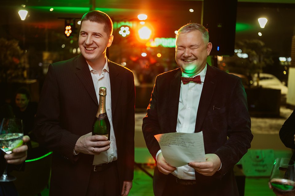 Two men in suits laughing at a celebration event, one holding a bottle of champagne and the other holding a paper, in a brightly lit indoor setting with green and red lighting.
