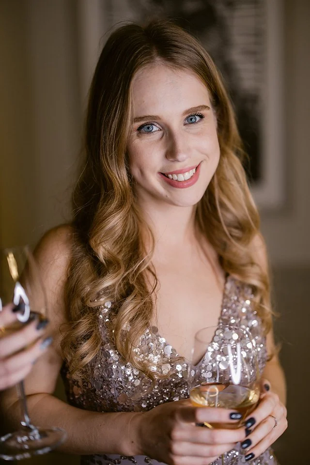 A young woman with long, wavy, light brown hair and blue eyes smiling while holding a glass of white wine, wearing a sparkly silver dress, at a social gathering or celebration.