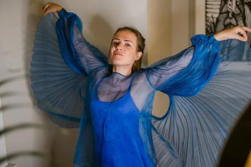 Woman in a sheer blue dress with pleated, flowing sleeves raising her arms.