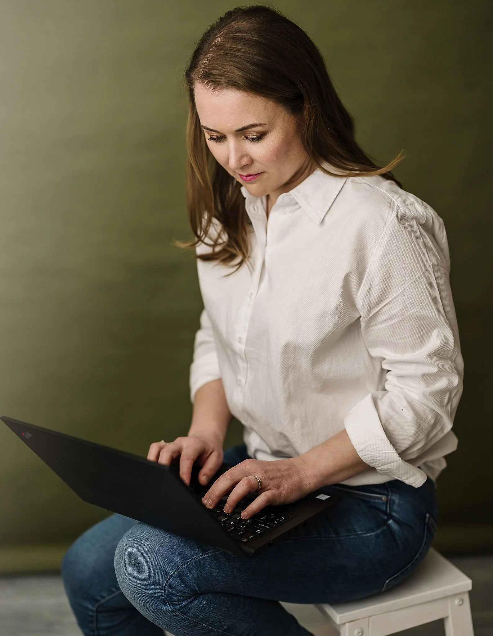 A woman sitting on a white chair, using a black laptop, wearing a white button-down shirt and blue jeans, with a green background.