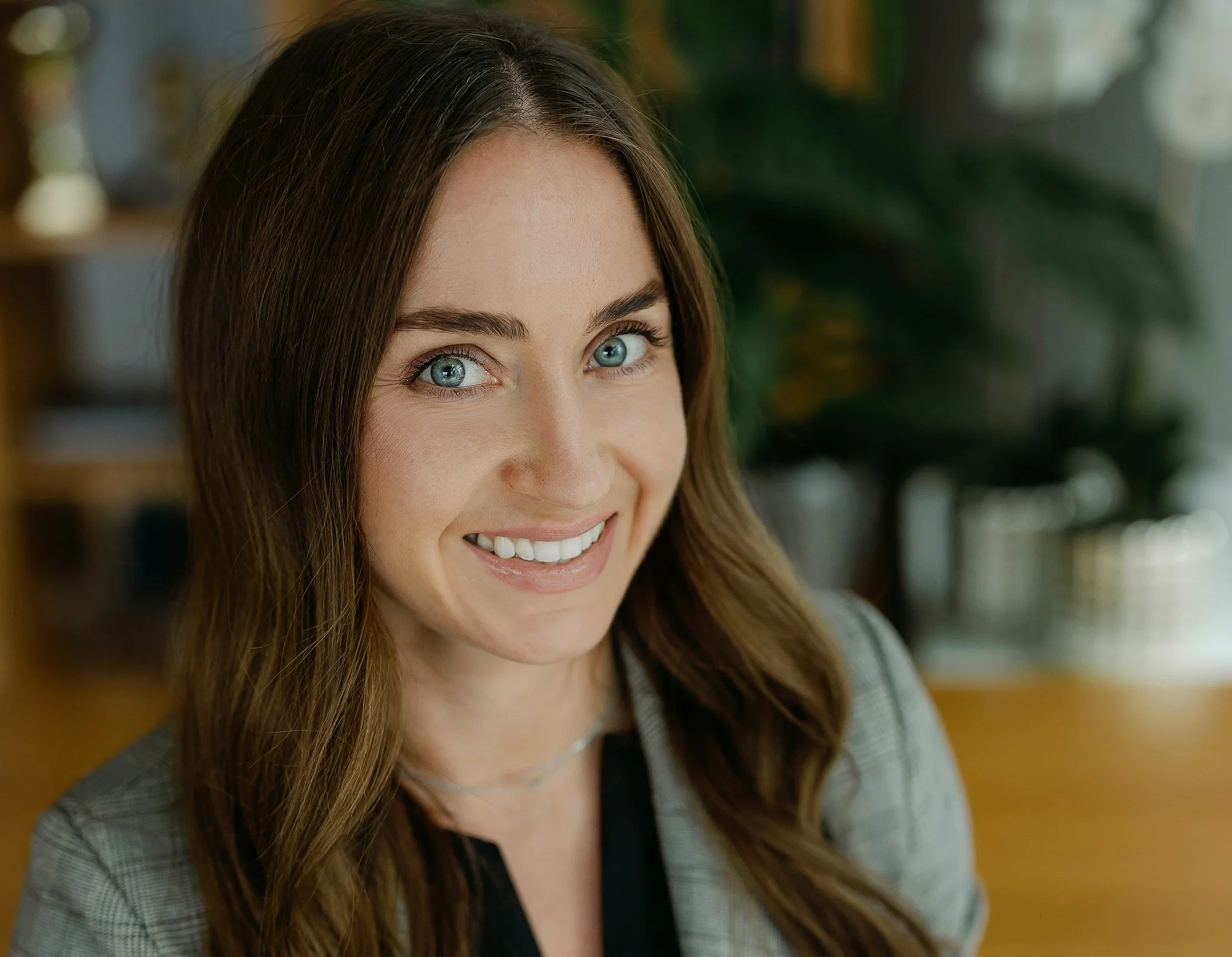 A smiling woman with long brown hair and blue eyes, wearing a gray blazer and black top, in an indoor setting with blurred greenery and furniture in the background.