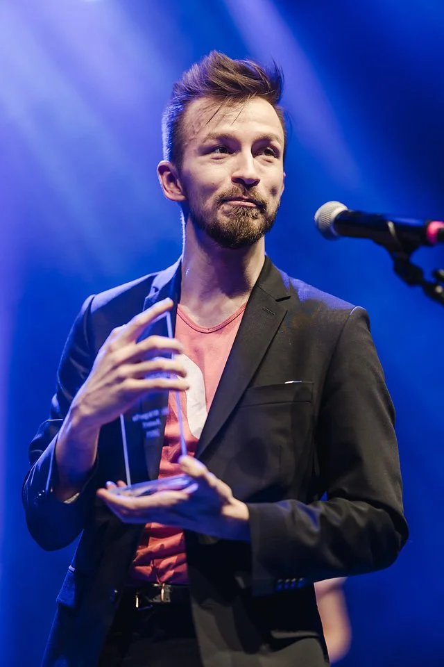 A man with a beard and styled hair standing on stage, holding an award or plaque, in front of a microphone, with blue stage lighting.