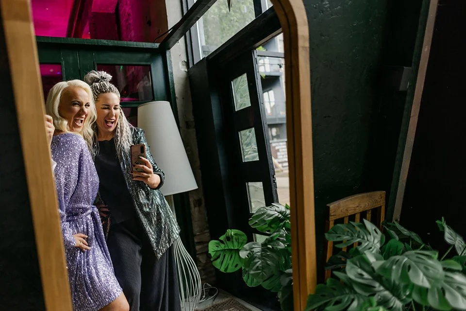 Two women are smiling and taking a selfie in front of a mirror, with green plants and a door with windows in the background.