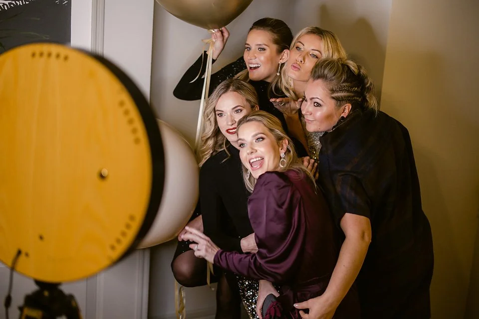 A group of five women taking a fun, candid photo together at a party. They are smiling and posing with balloons, standing close to a wall.