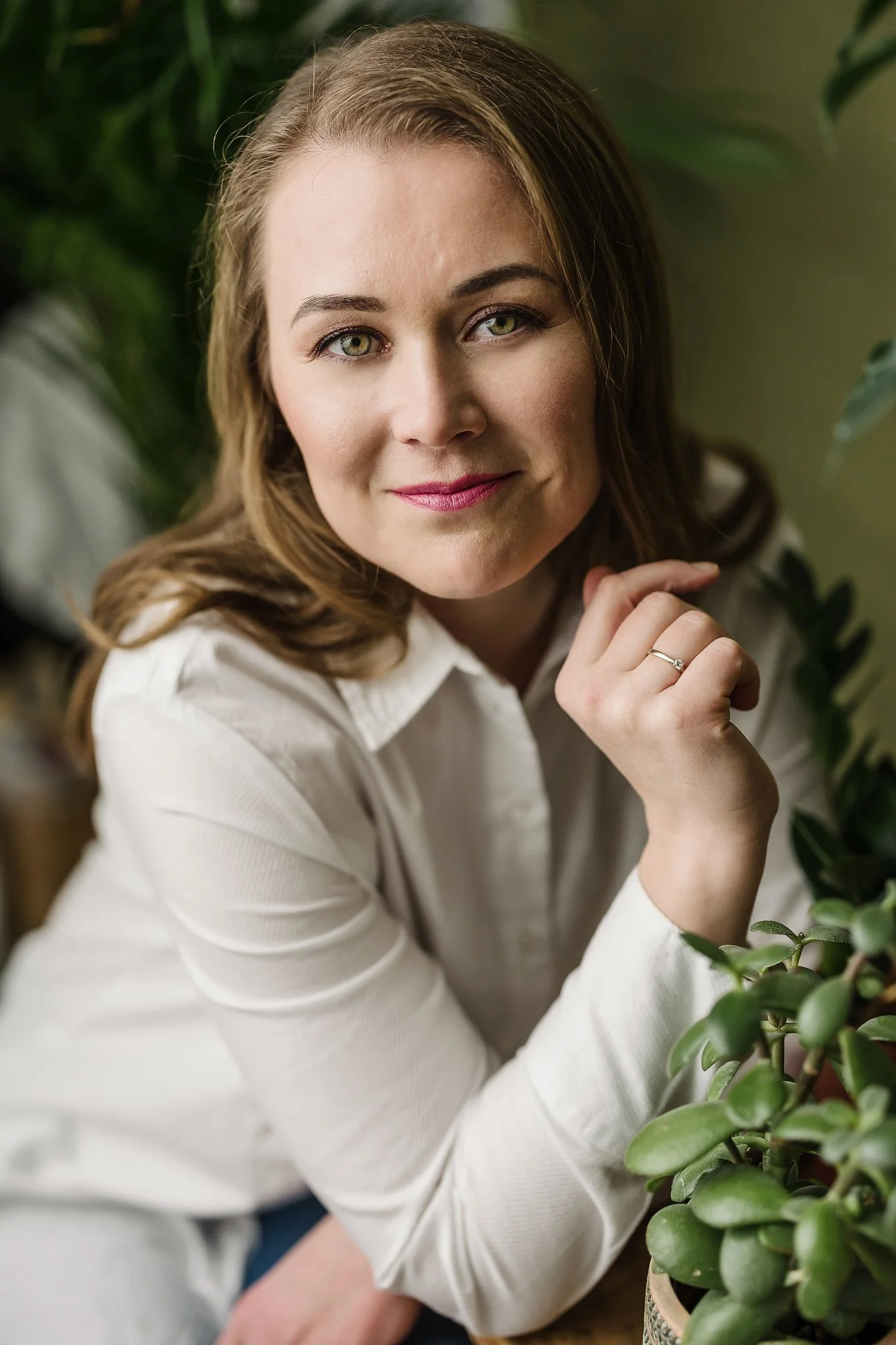 Close-up portrait of a woman with light brown hair, wearing a white shirt, sitting near green plants, smiling softly, with her chin resting on her hand and a ring on her finger.