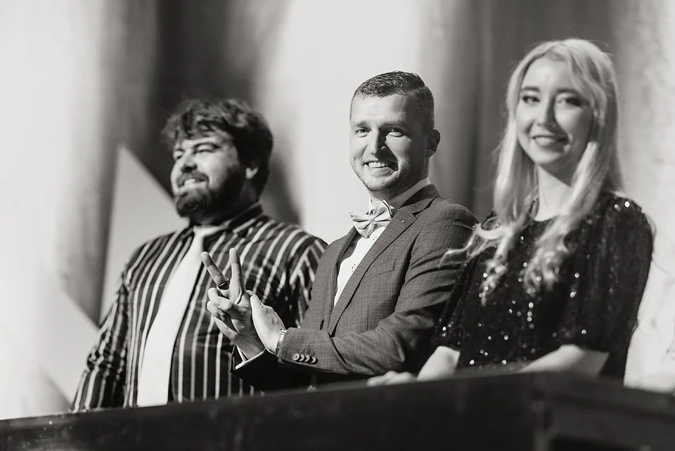 Three people, two men and a woman, smiling and seated at a table in a black and white photo, with the man in the middle making a peace sign.