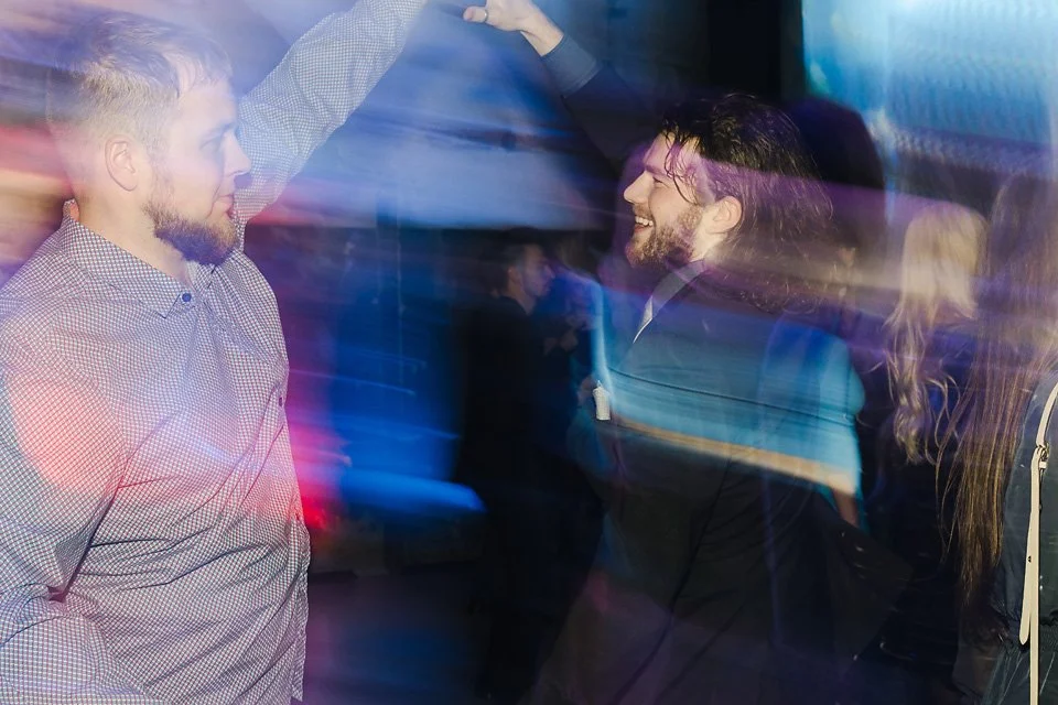 Two men dancing and smiling at a party with colorful light streaks.