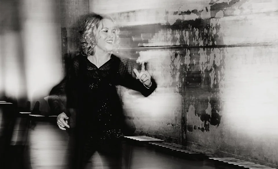 A woman smiling and pointing at a wall with abstract paint stains, standing near a piano in a room.