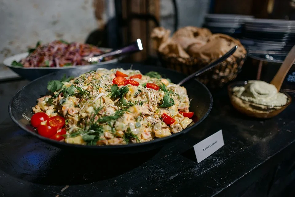 A large black bowl of potato salad garnished with cherry tomatoes and fresh herbs, with bowls of bread and dip in the background.