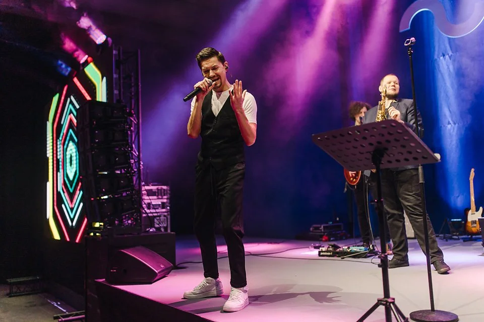 Performer singing into a microphone on stage with a band, colorful stage lighting, and musical instruments in the background.