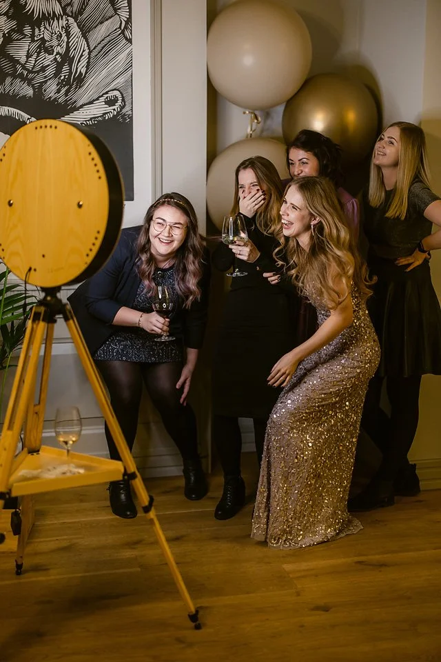 Group of five women at a party, laughing and looking at a photo booth camera. One woman is wearing a gold sequined dress, all holding wine glasses. Large balloons are in the background, and a photo booth setup is visible in the foreground.