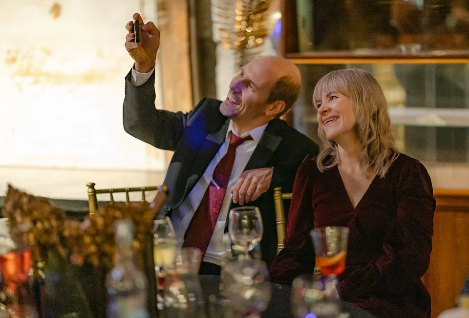 A man and woman sitting at a table, taking a selfie together at a social event or party.
