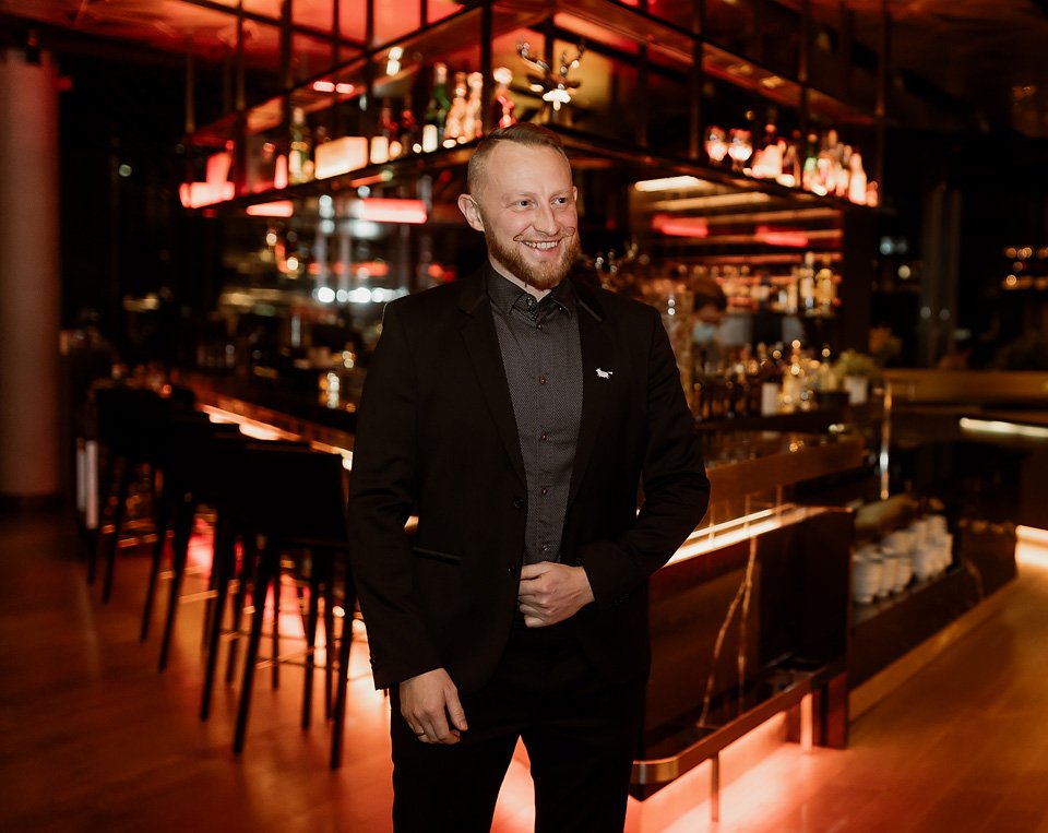A man in a black suit standing in a modern, dimly lit bar with a well-stocked counter and colorful lighting overhead.
